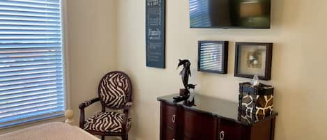 Un dormitorio con cama, una silla, cómoda, televisor y adornos en la pared.