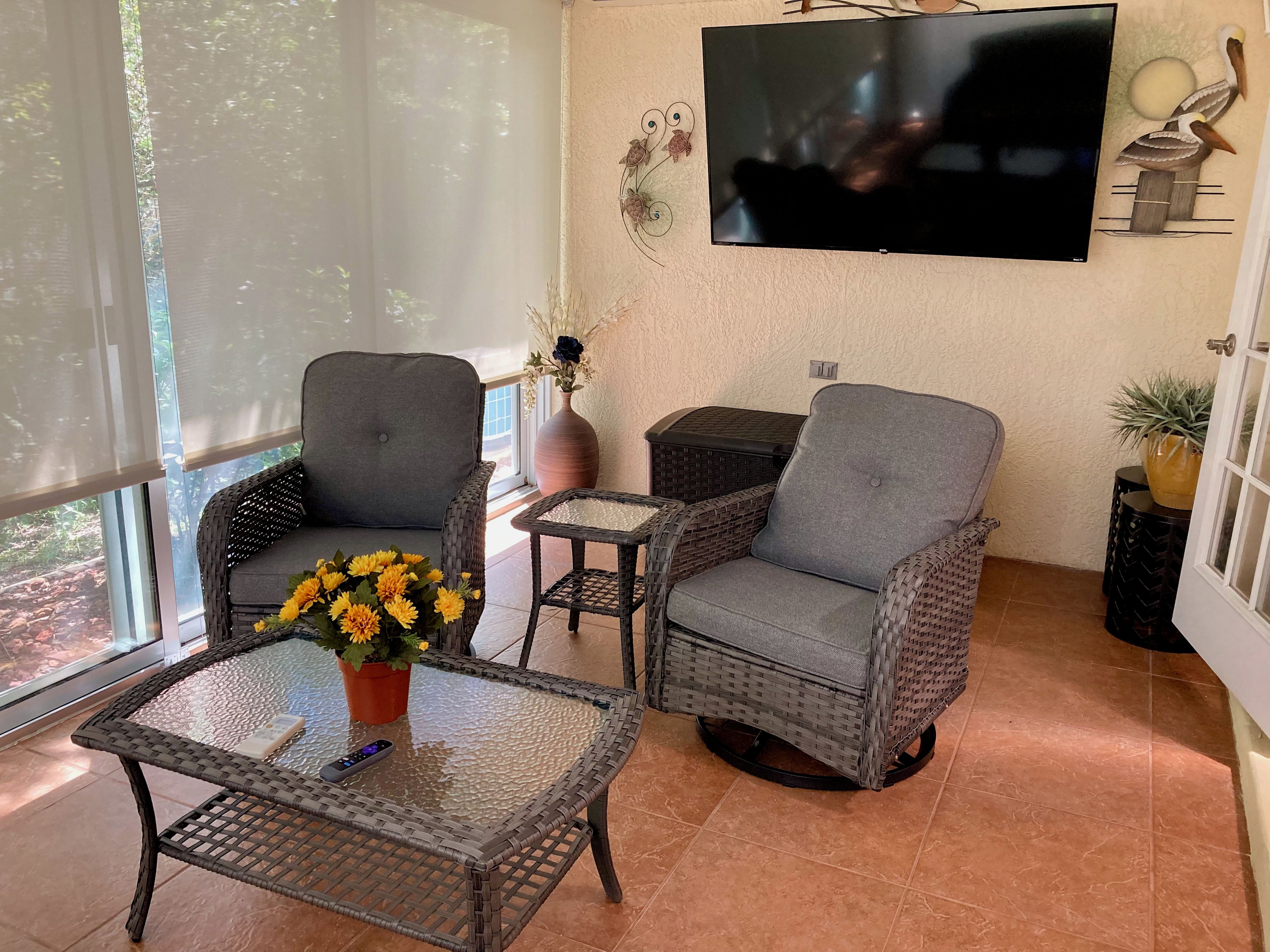 Ein Zimmer mit zwei grauen Korbstühlen, einem kleinen Tisch mit einer Vase voller gelber Blumen und einem an der Wand befestigten Fernseher.