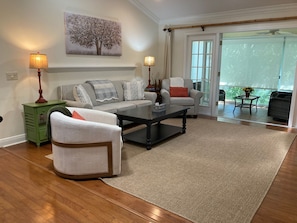 A living room with a sofa, armchair, coffee table, and lamps. There is a painting on the wall and a view through the glass door.