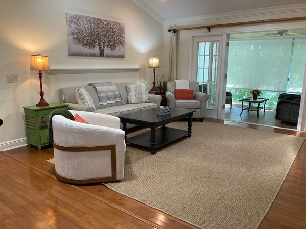 A living room with a sofa, armchair, coffee table, and lamps. There is a painting on the wall and a view through the glass door.