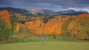 Un terrain de golf où deux joueurs évoluent, entourés d’un paysage automnal et de montagnes.