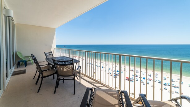 Een balkon met een tafel en stoelen, uitkijkend op een strand met parasols en mensen.