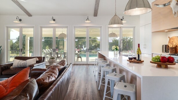 A spacious living area with a leather sofa, white bar stools, and a large window overlooking a pool.