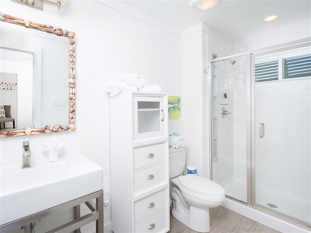 Une salle de bain avec un lavabo blanc, un miroir orné d’un cadre en forme de coquillage, des toilettes et une douche à parois de verre.
