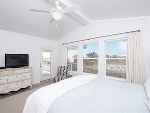 Ein Schlafzimmer mit einem großen Bett, einem Deckenventilator, einem Fernseher auf einer Kommode und Blick auf den Strand durch große Fenster.