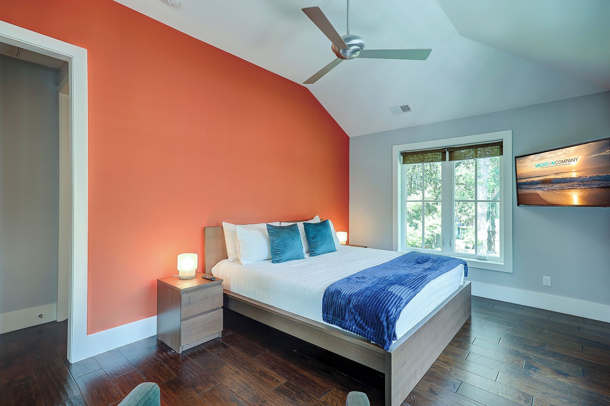 A bedroom with a bed, a nightstand, a ceiling fan, a window with a view, and a TV mounted on the wall.