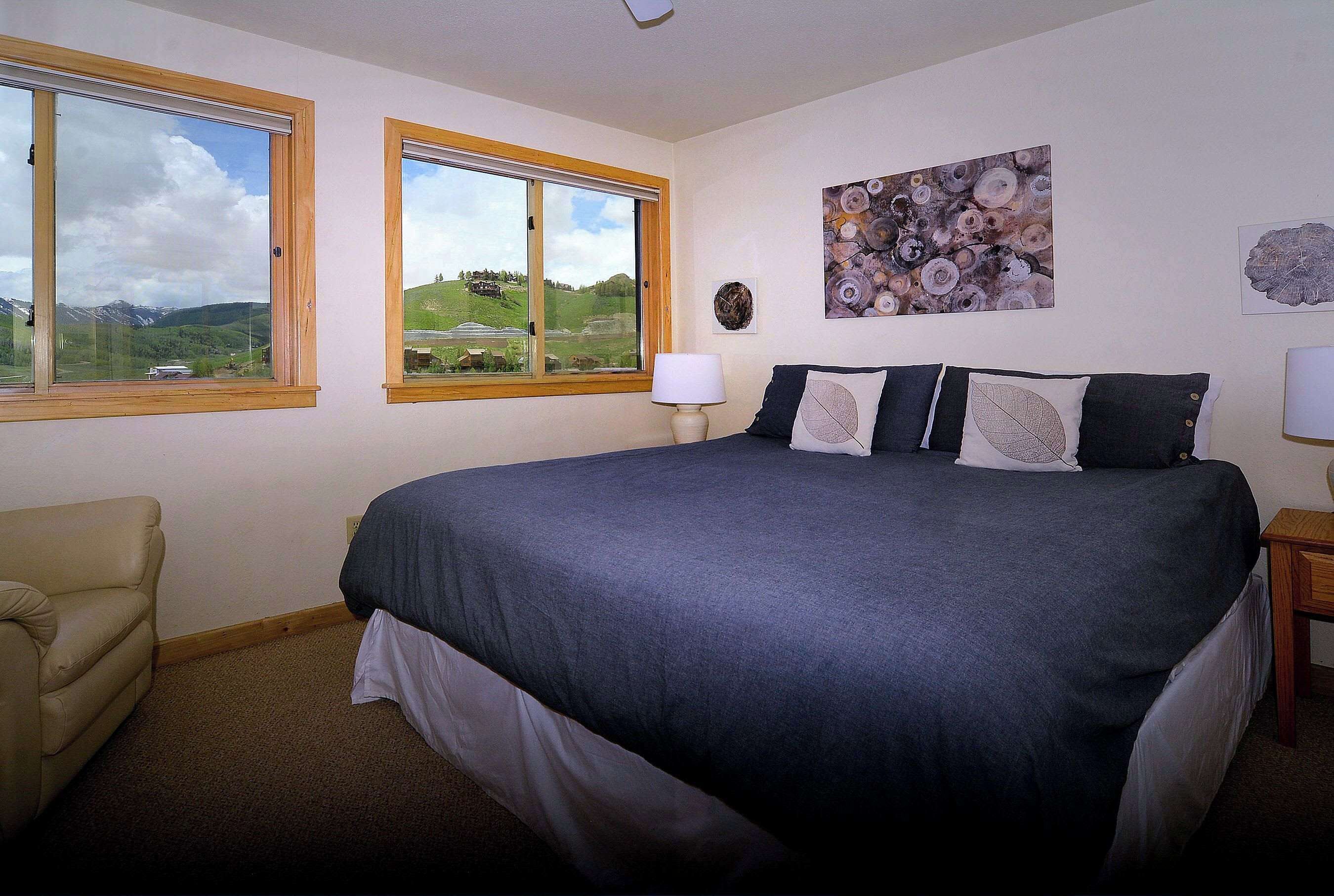 Une chambre à coucher avec un grand lit, deux fenêtres à cadres en bois, une chaise et des œuvres d’art accrochées au mur.