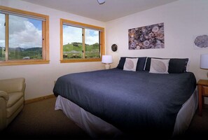 A bedroom with a large bed, two windows with wooden frames, a chair, and artwork on the wall. - Aspen Grove, Ski in/out, Hot Tub, Pet Friendly (Mt. Crested Butte)