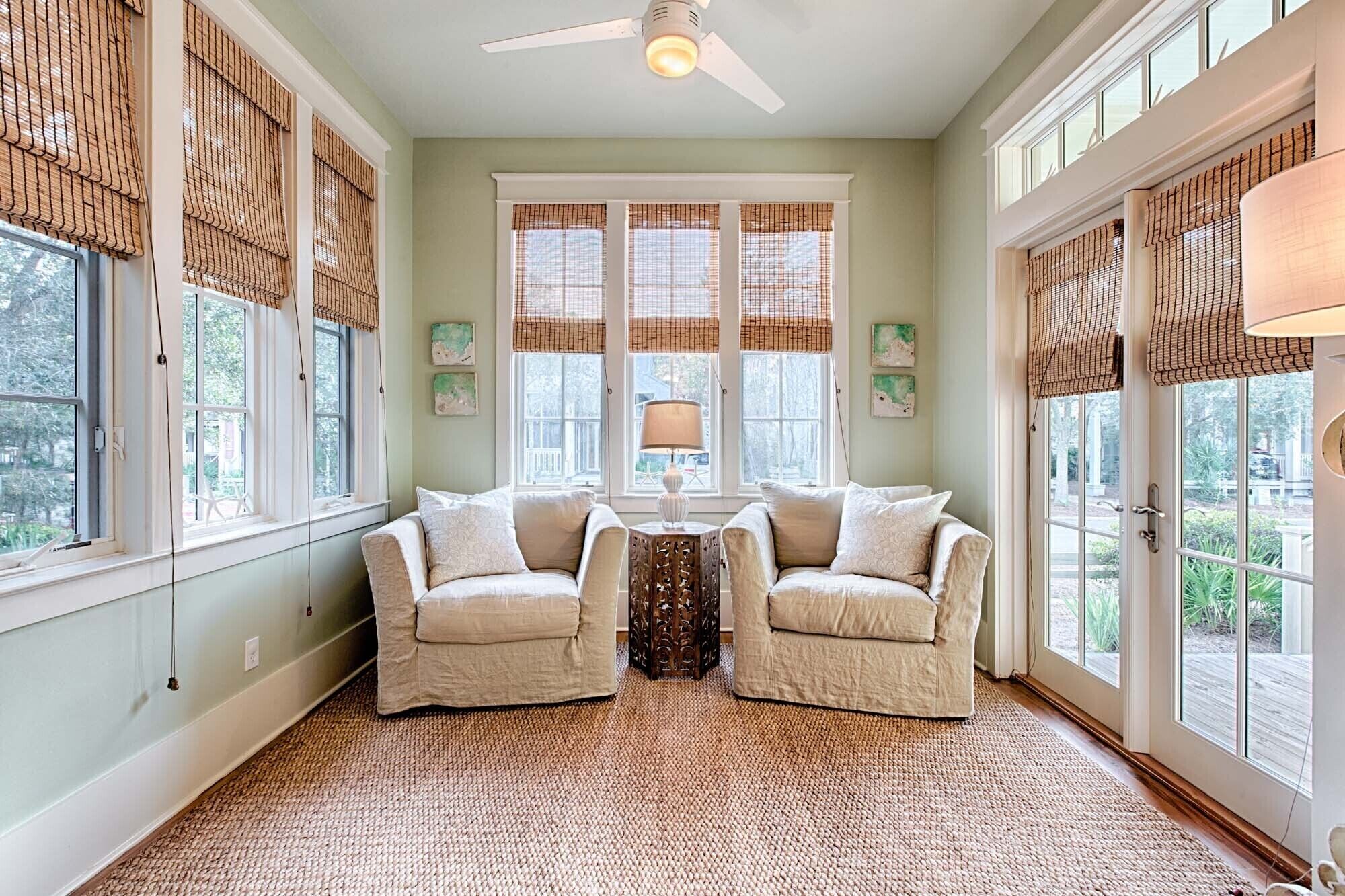 A room with two armchairs facing a small table, large windows with blinds, a ceiling fan, and a view of greenery outside.