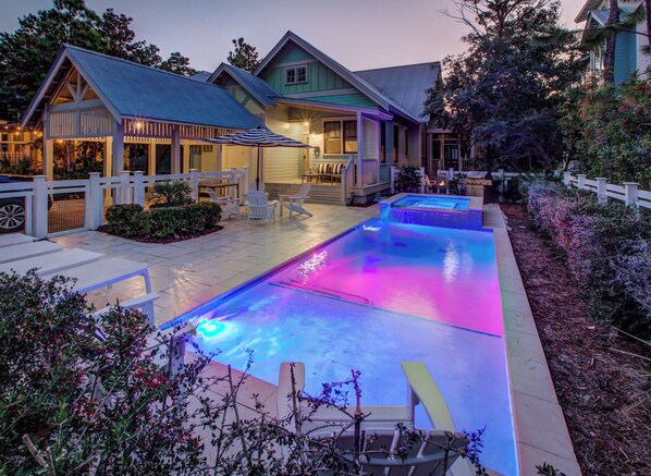 A swimming pool with a blue and pink light display, surrounded by outdoor seating and a house with a porch.