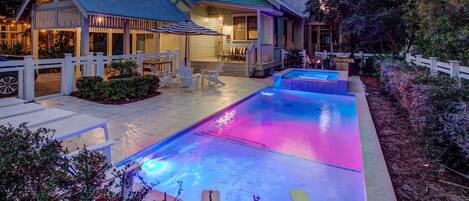 A swimming pool with a blue and pink light display, surrounded by outdoor seating and a house with a porch.
