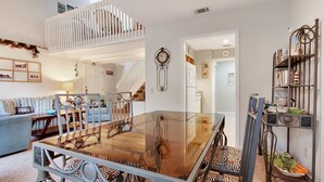 Un comedor amplio con una mesa de vidrio y sillas metálicas, una escalera que sube a un altillo y una estantería con diversos objetos.