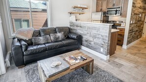 Una sala de estar moderna con un sofá de cuero negro, una mesa de centro de madera y una cocina con armarios de madera y un microondas.