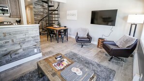 Una sala de estar moderna con una mesa de centro de madera, un sofá de cuero negro, un televisor montado en la pared y una escalera que conduce a un nivel superior.