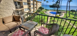 A balcony with a sofa, wicker chairs, and a table. It overlooks a pool area with a view of the ocean.