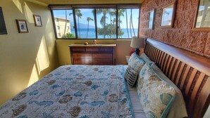 Quarto com cama, cômoda e vista para palmeiras e o oceano.