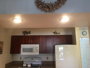 A kitchen with dark wooden cabinets, a white microwave, a stove, and a white refrigerator. There are two wall-mounted fish decorations and a clock on the wall. - Kihei Bay Vista #C-106 Best Location, Garden View, Cal King Bed, Hot Tub (Kihei)