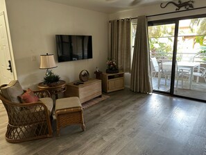 A living room with a flat-screen TV, wicker furniture, a small table with a lamp, and a sliding glass door leading to an outdoor area.