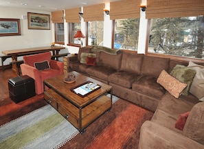 A cozy living room with a brown sofa, red armchair, and a wooden coffee table. There are framed pictures on the wall and a window with a view of trees.