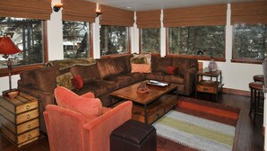 A cozy living room with a brown sofa, a red armchair, and a coffee table. There are large windows with blinds, a lamp, and a small table with a vase. - Perfect Views and Location in Lionshead! Well Appointed Condo Steps from Lifts (Vail)