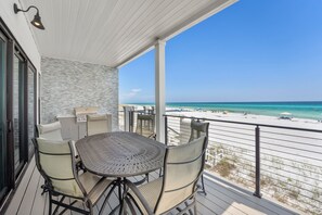 Uma área externa coberta com mesa, cadeiras, churrasqueira e vista para a praia.