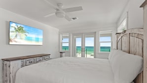Une chambre avec un grand lit, un ventilateur de plafond, un téléviseur diffusant une scène de plage et une vue sur l’océan par des portes ouvertes.
