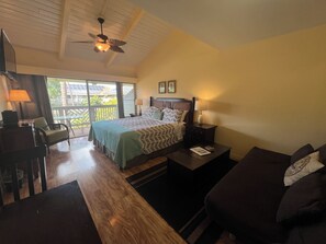 Een slaapkamer met een bed, een bureau, een stoel, een bank, een plafondventilator en een balkon met uitzicht op palmbomen.