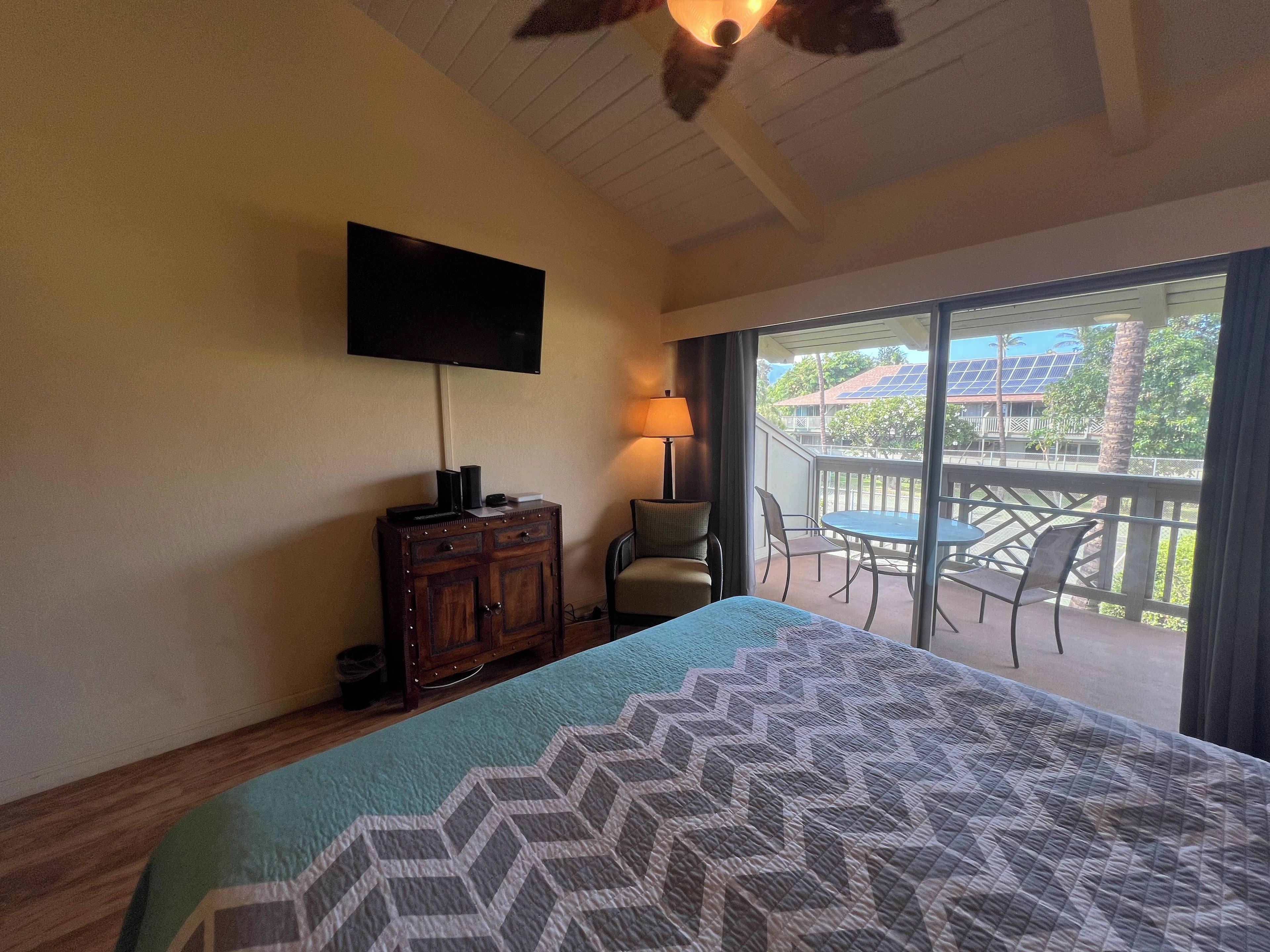 A hotel room with a bed, a TV mounted on the wall, a wooden cabinet, a chair, and a balcony with a table and chairs.