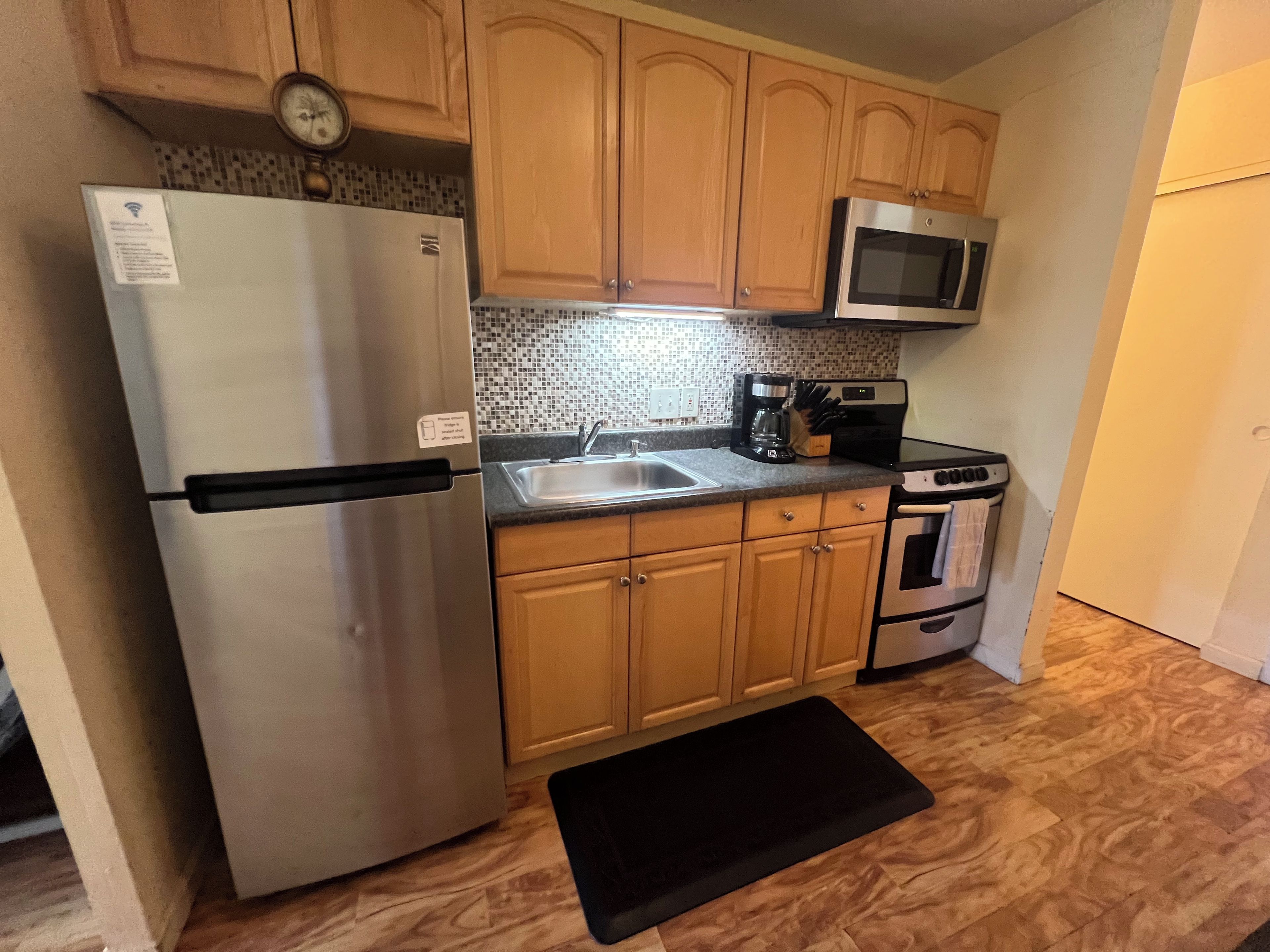 A compact kitchen with wooden cabinets, a stainless steel refrigerator, a microwave, a coffee maker, and a stove.