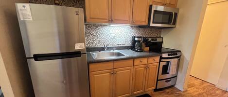 A compact kitchen with wooden cabinets, a stainless steel refrigerator, a microwave, a coffee maker, and a stove.