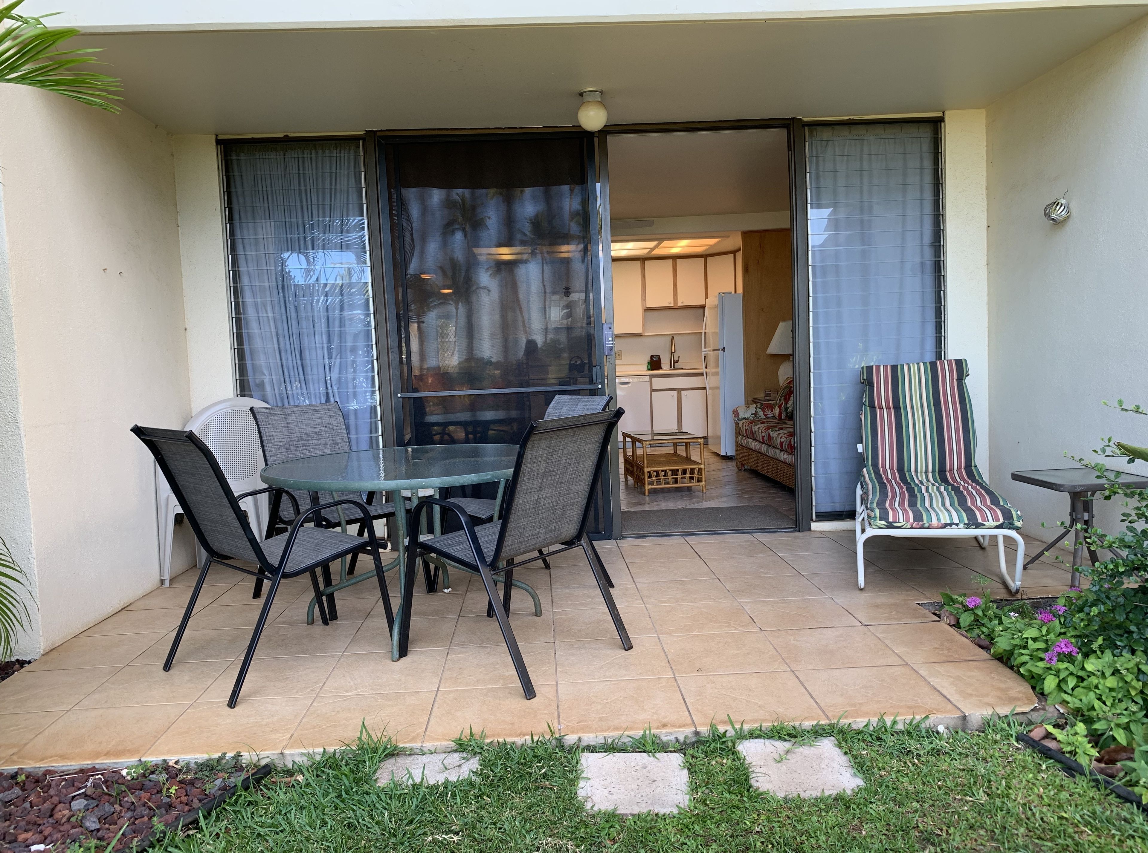 En patio med et glasbord og stole, en stribet liggestol og udsigt til et rum med et køkken og en sofa.