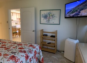 A hotel room with a bed, a TV mounted on the wall, a small wooden shelf, and a white air conditioner.