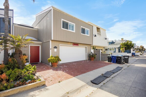A two-story residential building with a garage, a red door, and a driveway leading to the street.