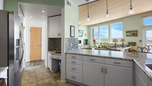 A kitchen with white cabinets, a granite countertop, and a view of the ocean through large windows. - 3441O Mollies by the Sea (Oxnard)
