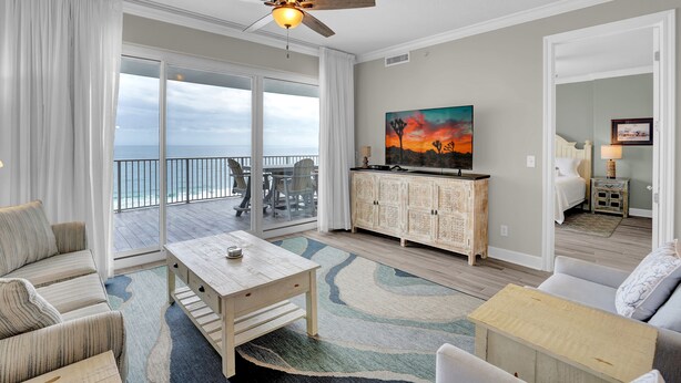 A spacious living room with a large sliding glass door leading to a balcony, a ceiling fan, a dining area, and a view of the ocean. - Regency Isle 911 by Liquid Life (Orange Beach)