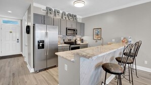 A kitchen with a stainless steel refrigerator, a microwave, and a stove. There is a bar area with bar stools. The word "BEACH" is visible on the wall. - Regency Isle 911 by Liquid Life (Orange Beach)