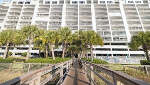 A multi-story hotel with a central walkway, surrounded by palm trees and a wooden fence. - Regency Isle 911 by Liquid Life (Orange Beach)