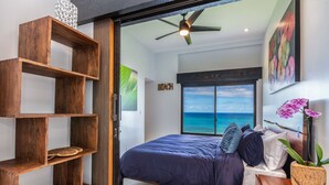 A bedroom with a bed, wooden shelves, a ceiling fan, a view of the ocean, and a beach-themed wall art. - SeaLodge A-7 "Honu Hale" Spectacular Ocean Views! (Princeville)