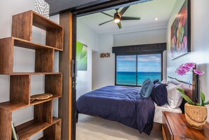 Enjoy ocean views from the large window in this serene bedroom featuring a comfortable bed and stylish shelving unit.