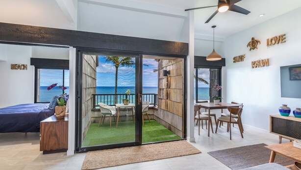 A modern living room with a large sliding glass door leading to a balcony with a view of the ocean. There is a dining area with a table and chairs, and a TV mounted on the wall. - SeaLodge A-7 "Honu Hale" Spectacular Ocean Views! (Princeville)