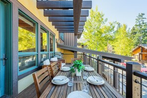 Um deck de madeira com uma mesa posta para quatro pessoas, área de estar ao ar livre e vista para árvores e um edifício ao fundo.