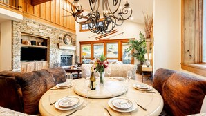 A dining area with a round table set for four, surrounded by leather sofas, a stone fireplace, and a large chandelier. - Pristine Mountain Home. Perfect for Families - Hot Tub, Media Room & Elevator (Park City)