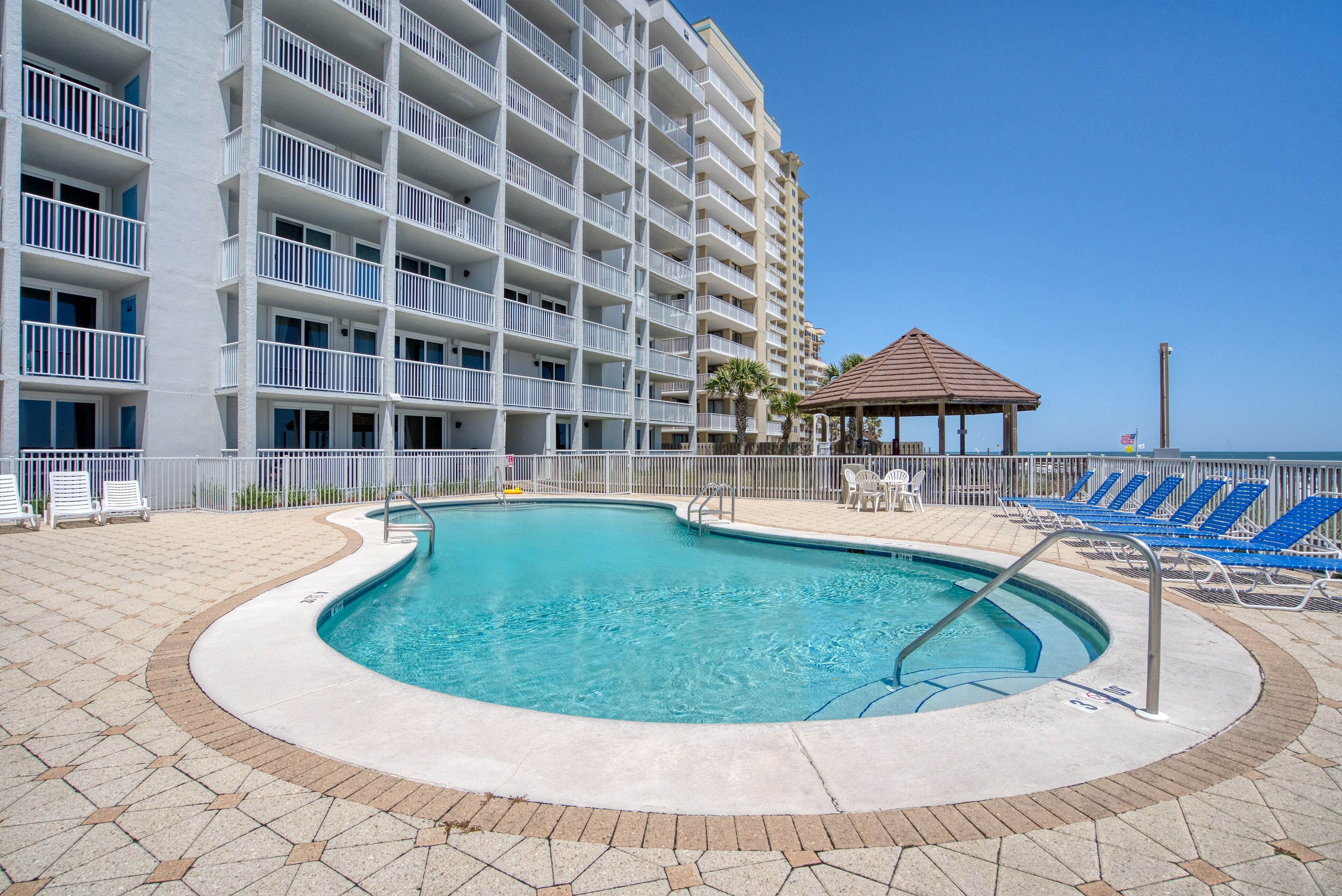 A hotel pool area with a clear blue pool, white balconies, and blue lounge chairs.