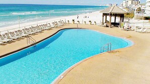 Una piscina con bordo curvo, adiacente a una spiaggia con lettini e un padiglione.