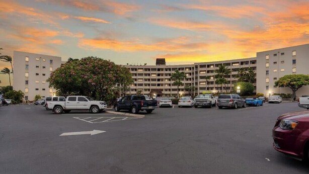 A parking lot with multiple cars, a large building with a flat roof, and a sunset sky. - Your Refreshing Escape by the Ocean! Marvelous Views - Royal Mauian 209 (Kihei, Maui)