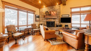 A cozy living room with a stone fireplace, wooden furniture, and large windows offering a view of the mountains. - Modern Mountain Design, High-End Kitchen, Theater Room & Hot Tub! (Park City)