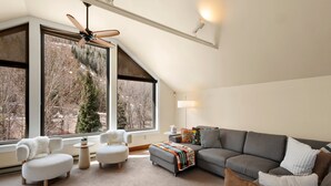 Ein modernes Wohnzimmer mit Deckenventilator, großen Fenstern, einem grauen Sofa, weißen Sesseln und einem kleinen Tisch mit Pflanze.