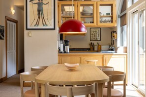 A dining area with a wooden table and chairs, a red pendant light, and a kitchen in the background.