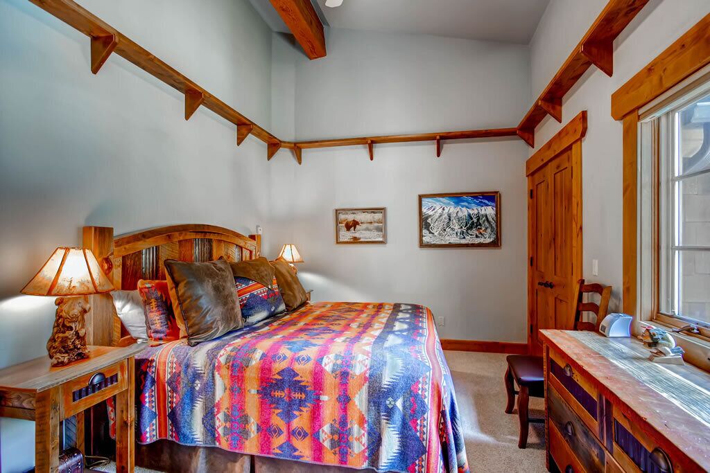 A bedroom with a wooden headboard, a colorful bedspread, a desk with a chair, and a window with a view of the mountains.