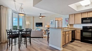 A modern kitchen with wooden cabinets, a central island, and a dining area with chairs and a table. - Paradise at the Palms * Summer Specials! Book Now! 2 King Beds~2 Full Baths! (Destin)